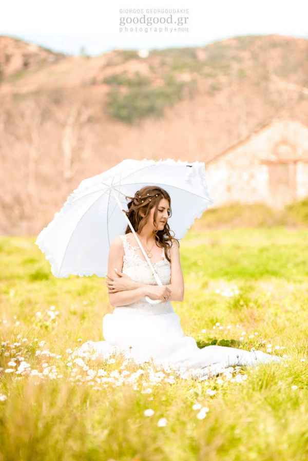 Μία νύφη κάθεται στο χορτάρι και κρατάει μία λευκή ομπρέλα σε φωτογράφιση επόμενης μέρας γάμου